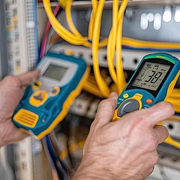 Устройство для измерения электрических параметров с красными щупами, подключенное к электрической панели