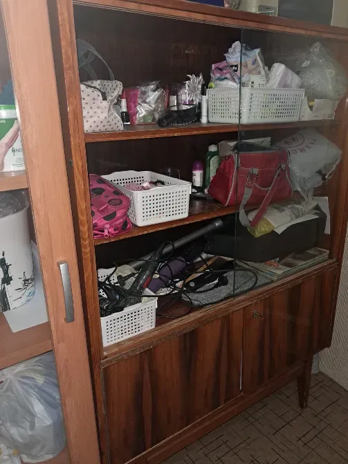 Деревянный шкаф с витриной, заполненный различными косметическими средствами, аксессуарами и бытовыми предметами. Внутри видны корзины и пакеты с изделиями