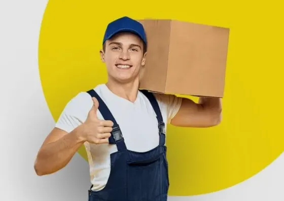 Молодой человек в рабочем комбинезоне и голубой кепке держит коробку на плече и показывает большой палец вверх на ярком фоне