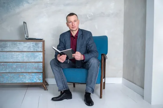 Мужчина сидит на синем кресле с книгой в руке, рядом с ним стоит стол и ноутбук