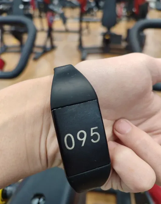Чёрный фитнес-браслет с номером 095 на руке на фоне тренажёрного зала