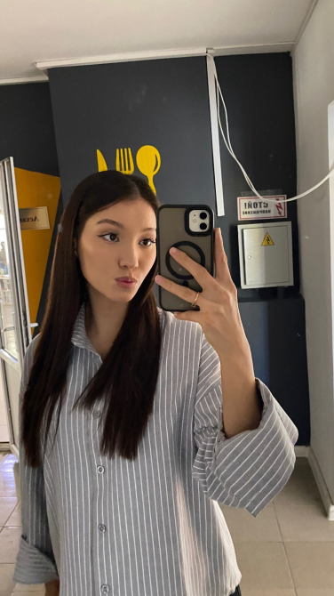 Молодая женщина с длинными волосами в полосатой рубашке, делающая селфи в помещении с темной стеной и графикой на стене