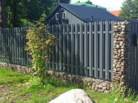 Серая металлическая ограда с вертикальными планками на фоне домов и зелени