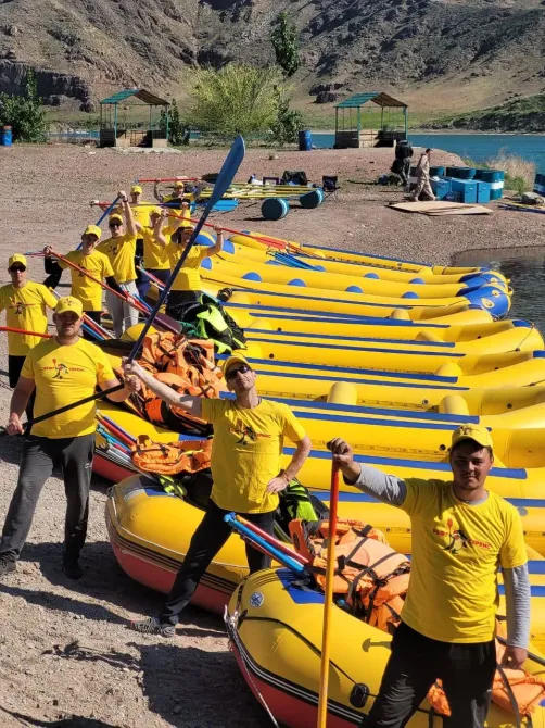 Группа людей в желтых футболках позирует с веслами рядом с рядами рафтов на берегу реки