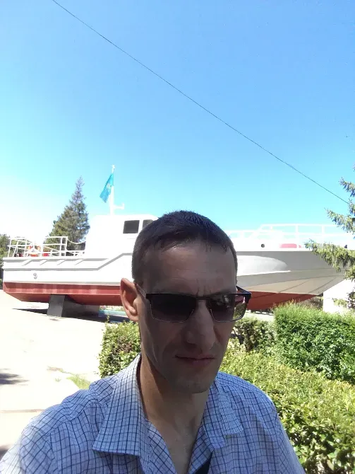 Күн көзілдірігін тағып тұрған адам қайықтың жанында, ал фондағы ашық көк аспан.