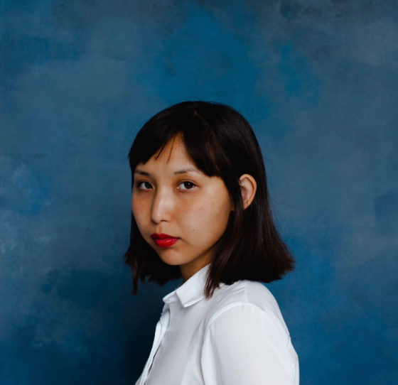 Женщина с короткими черными волосами, в белой рубашке на фоне синей стены, смотрит в камеру