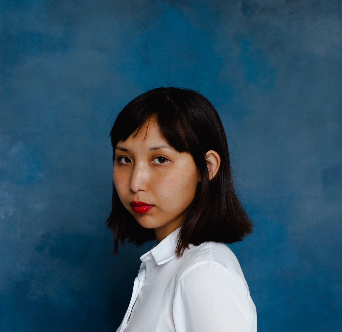 Женщина с короткими черными волосами, в белой рубашке на фоне синей стены, смотрит в камеру