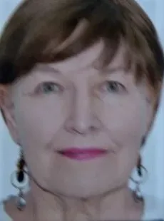 Портрет пожилой женщины с короткими каштановыми волосами и серьгами. Она смотрит прямо в камеру. На фоне размытая светлая поверхность