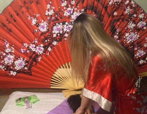 Женщина в красном кимоно с длинными светлыми волосами, стоящая перед большым декоративным веером, украшенным цветами сакуры, на фоне малиновой стены. 