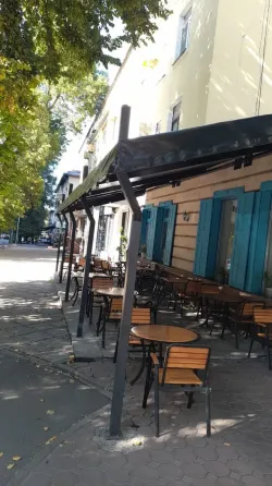 Көшедегі кафеде бос үстелдер, айналасында ағаштар мен ғимараттар.