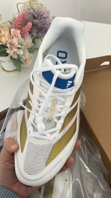 Белые кроссовки с золотыми полосами, удерживаемые в руке, с видимым логотипом на язычке