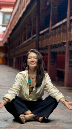 Женщина в светлом свитере и темных брюках, сидит на ступенях с красной стеной на фоне