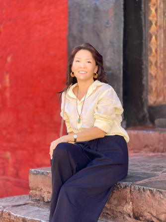 Женщина в светлом свитере и темных брюках, сидит на ступенях с красной стеной на фоне