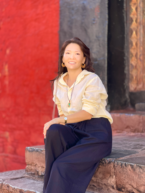 Женщина в светлом свитере и темных брюках, сидит на ступенях с красной стеной на фоне