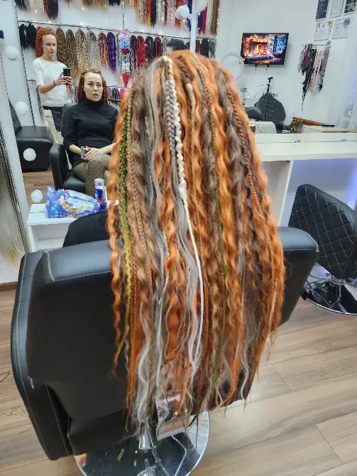 Прическа с длинными косами в оранжево-зеленых оттенках, сидящая на кресле в салоне