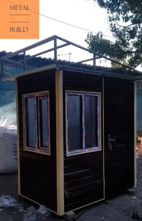 Металлическая постройка с крышей, окнами и дверью. Строение выполнено из темного металла, расположено на улице, окружено зелеными деревьями