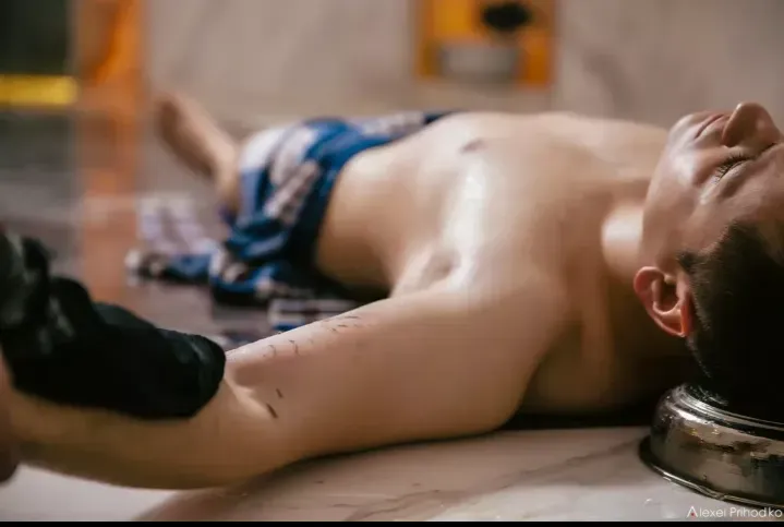 Молодой человек лежит на поверхности, его тело частично обнажено, он расслаблен и смотрит вверх. Вокруг него видны детали интерьера