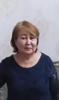 Женщина средних лет с короткой светлой прической, одетая в темную блузку, стоит на фоне стен с небольшими потертостями