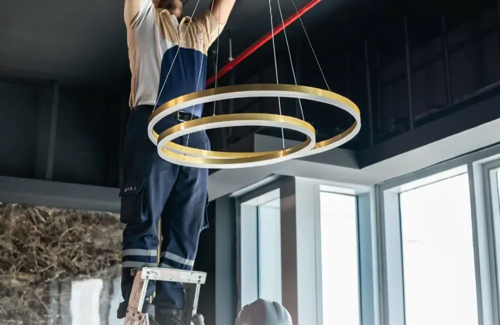Рабочий устанавливает современный светильник в интерьере с большими окнами; другой рабочий находится внизу на лестнице