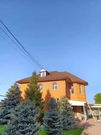 Екі қабатты, мансардалы, дөңгелек терезелері бар қызғылт сары үй, ашық көк аспан фонында. Алдыңғы жоспарында жасыл шыршалар көрінеді.