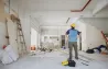 Каска киген жұмысшы бос бөлмедегі қабырғалары бұзылған және құрылыс материалдарымен толы жөндеу процесін бағалауда