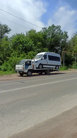 Грузовик с прицепом, на котором находится фургон, стоит на обочине дороги рядом с зелеными деревьями