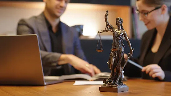 Офистегі Фемида мүсіні, үстел үстінде салмағы бар, құжаттармен және ноутбукпен жұмыс істеп отырған адамдар.