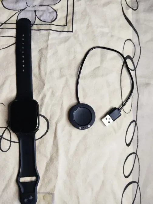 Чёрные часы на ремешке и зарядное устройство с USB, размещённые на светлом фоне с узором
