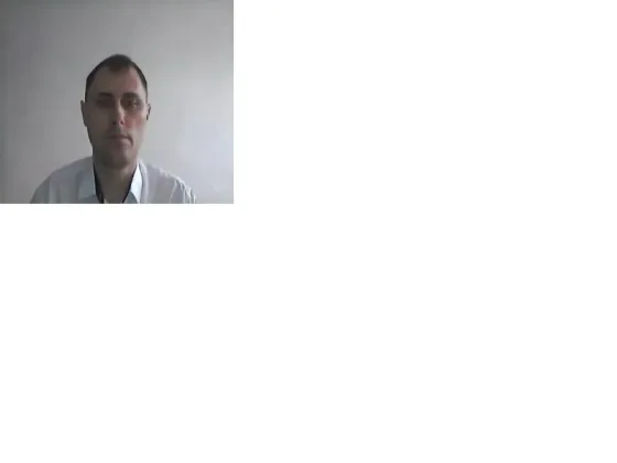 Ақ фондағы адам, жейде мен аксессуарлар киіп, камера алдында отырады.