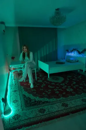 Ақ төсек пен жасыл жарығымен бөлме, қыз орындықта отыр