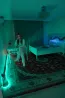 Ақ төсек пен жасыл жарығымен бөлме, қыз орындықта отыр