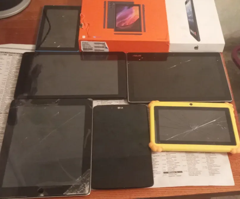 Планшеты различной модели и цвета на столе, включая коробки от iPad и других моделей, с видимыми трещинами на некоторых экранах