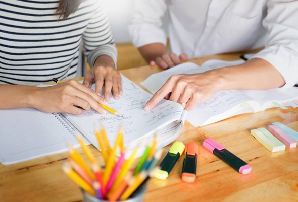 Два человека работают над учебными материалами за столом, пишут и рисуют в тетради, рядом с ними стоят яркие фломастеры и карандаши